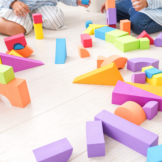 Bunte Schaumstoffbausteine auf hellem Holzboden, Kinder spielen im Hintergrund. Weniger Spielzeug ist manchmal mehr. q